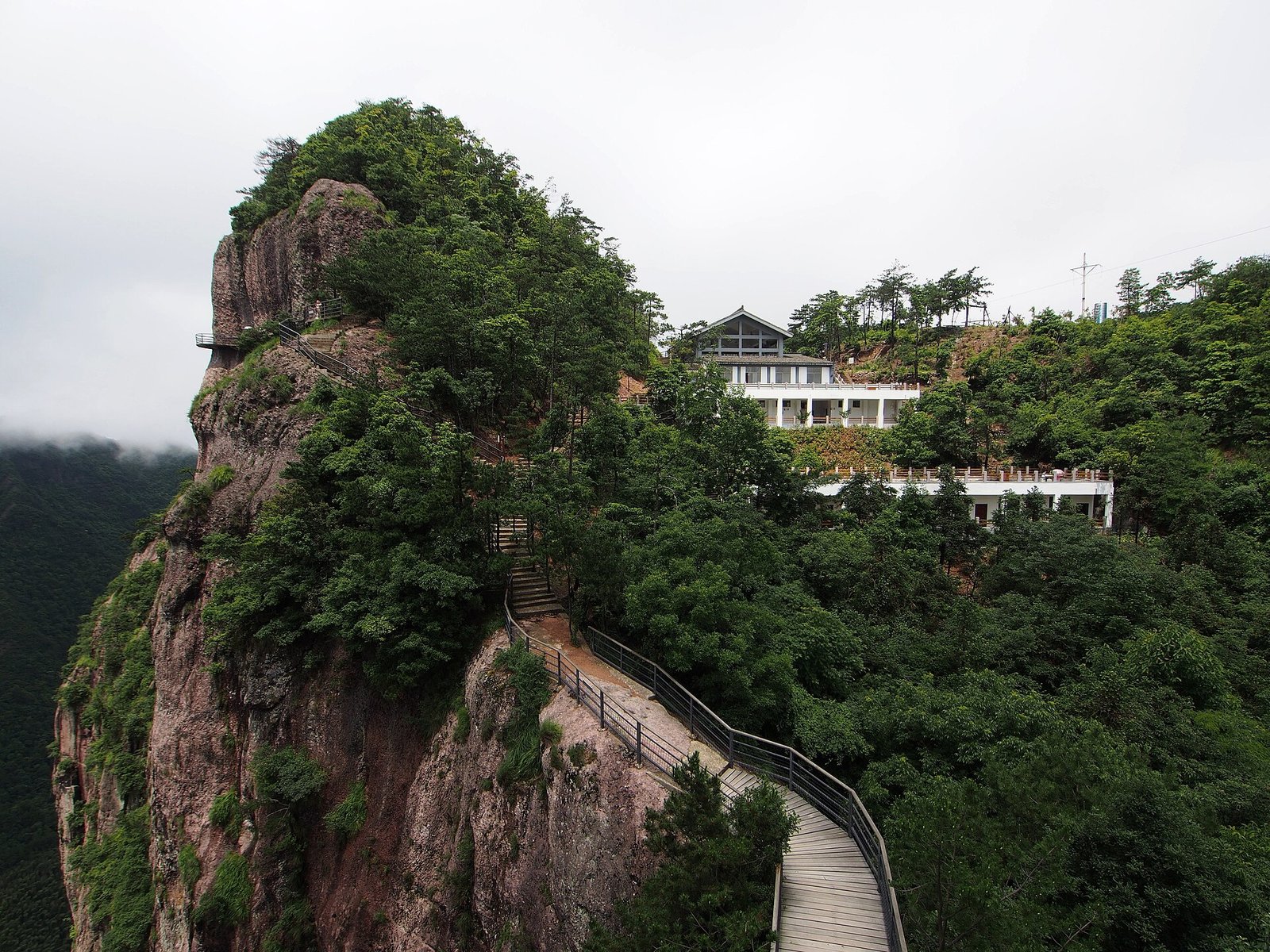 A rhyolite pillar wrapped in forest at Shenxianju scenic area in Xianju, Zhejiang, with a cliff walkway cut into the rock and cloud sitting in the valley behind