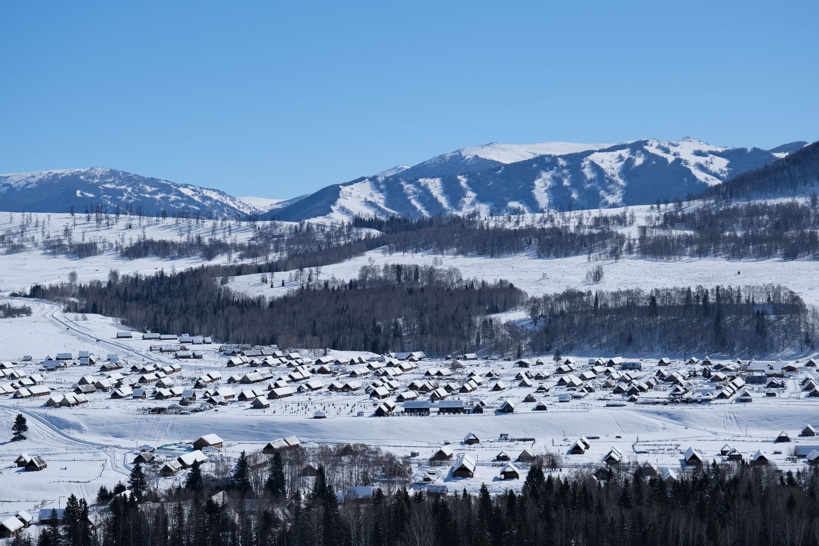 The snow-covered wooden houses of Hemu village in Altay, Xinjiang, with forested mountains rising behind