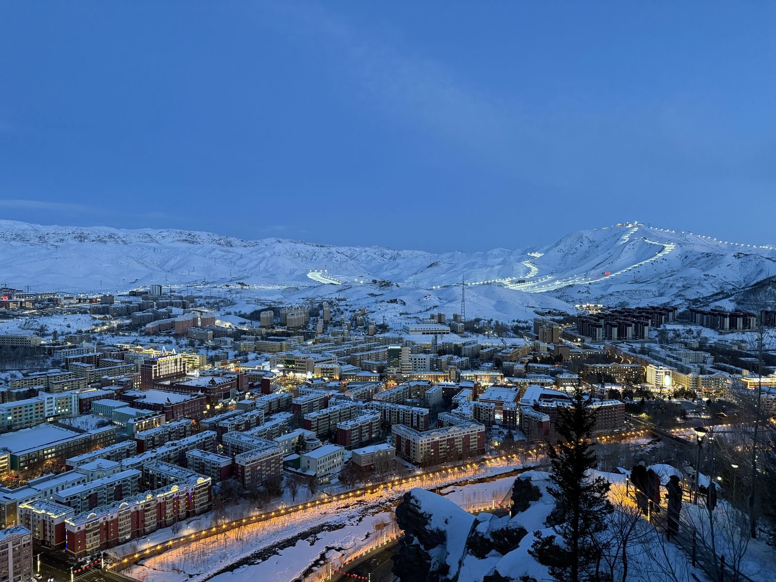 The city of Altay at dusk in winter, with the floodlit ski runs of Jiangjunshan ski resort lit up on the mountain above