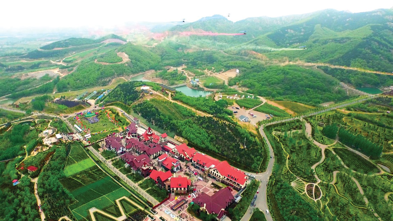 An aerial view of a green valley containing a cluster of red-roofed European-style buildings, roads, a small lake and terraced fields