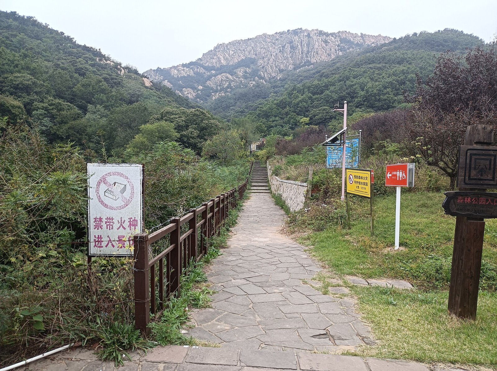 A stone-paved path leading between trees towards green hills, with a low entrance structure and signage