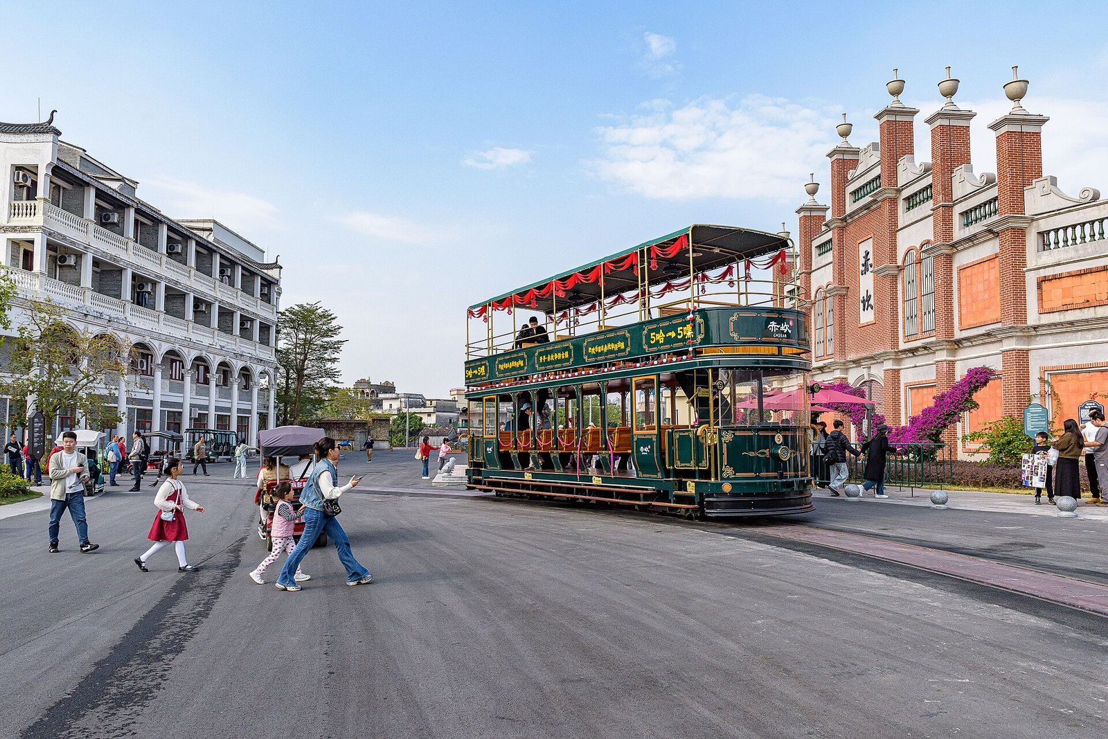 A green and cream open-sided tram standing at a stop in front of arcaded buildings, with passengers boarding
