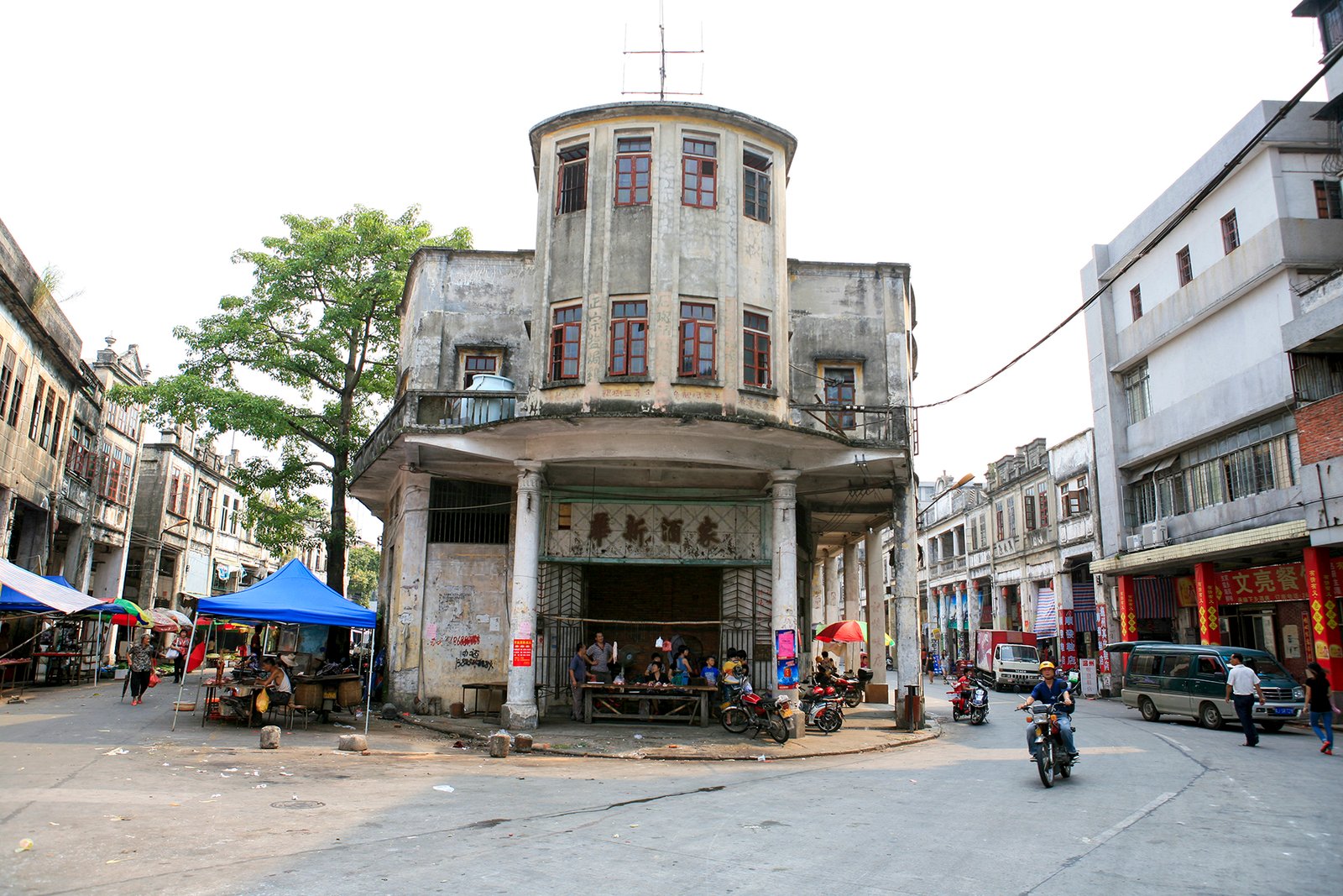 A street corner in Chikan with a curved corner building, market stalls under blue tarpaulins and motorbikes parked in the road