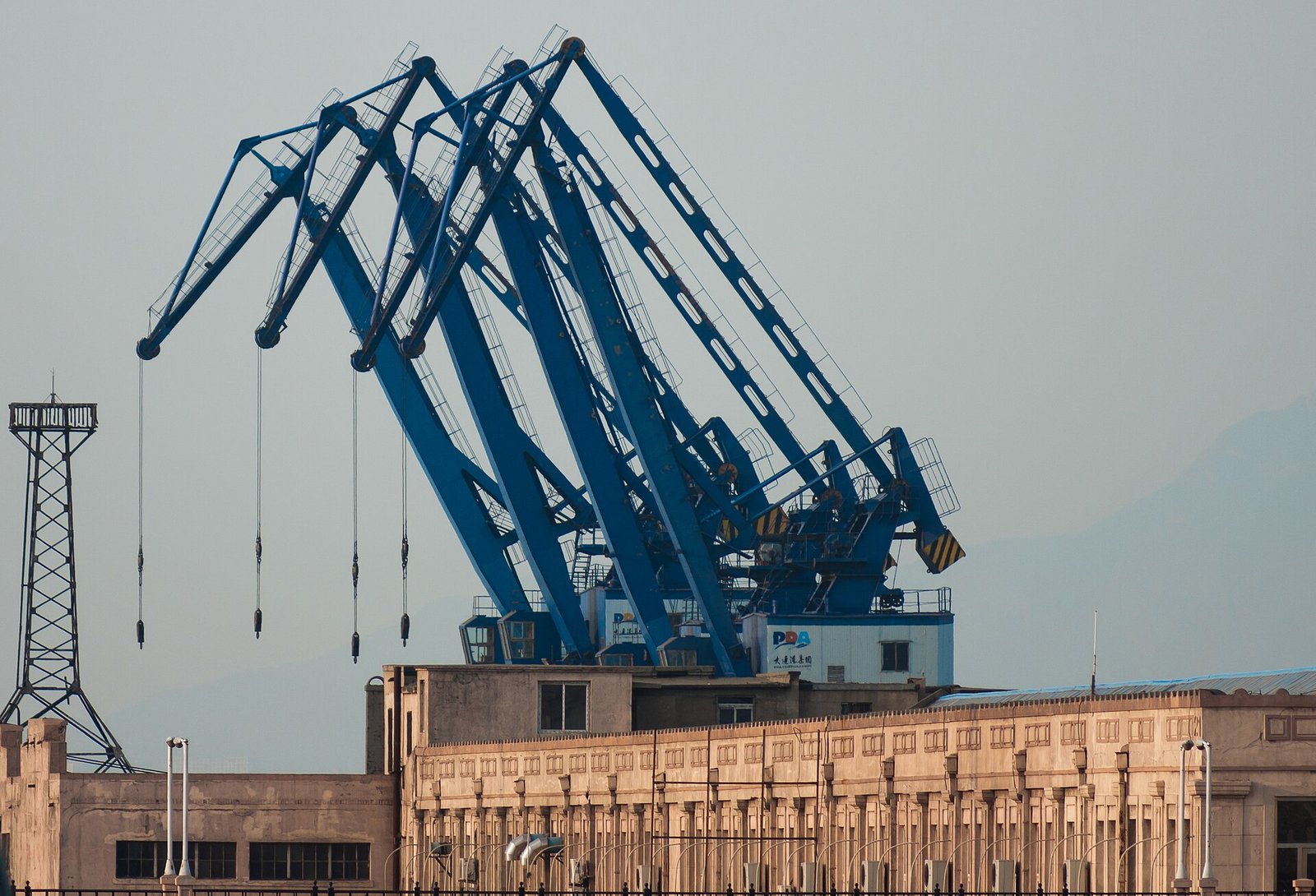 Blue steel crane booms over a dock wall in Dalian harbour, with a lattice tower and low industrial buildings behind