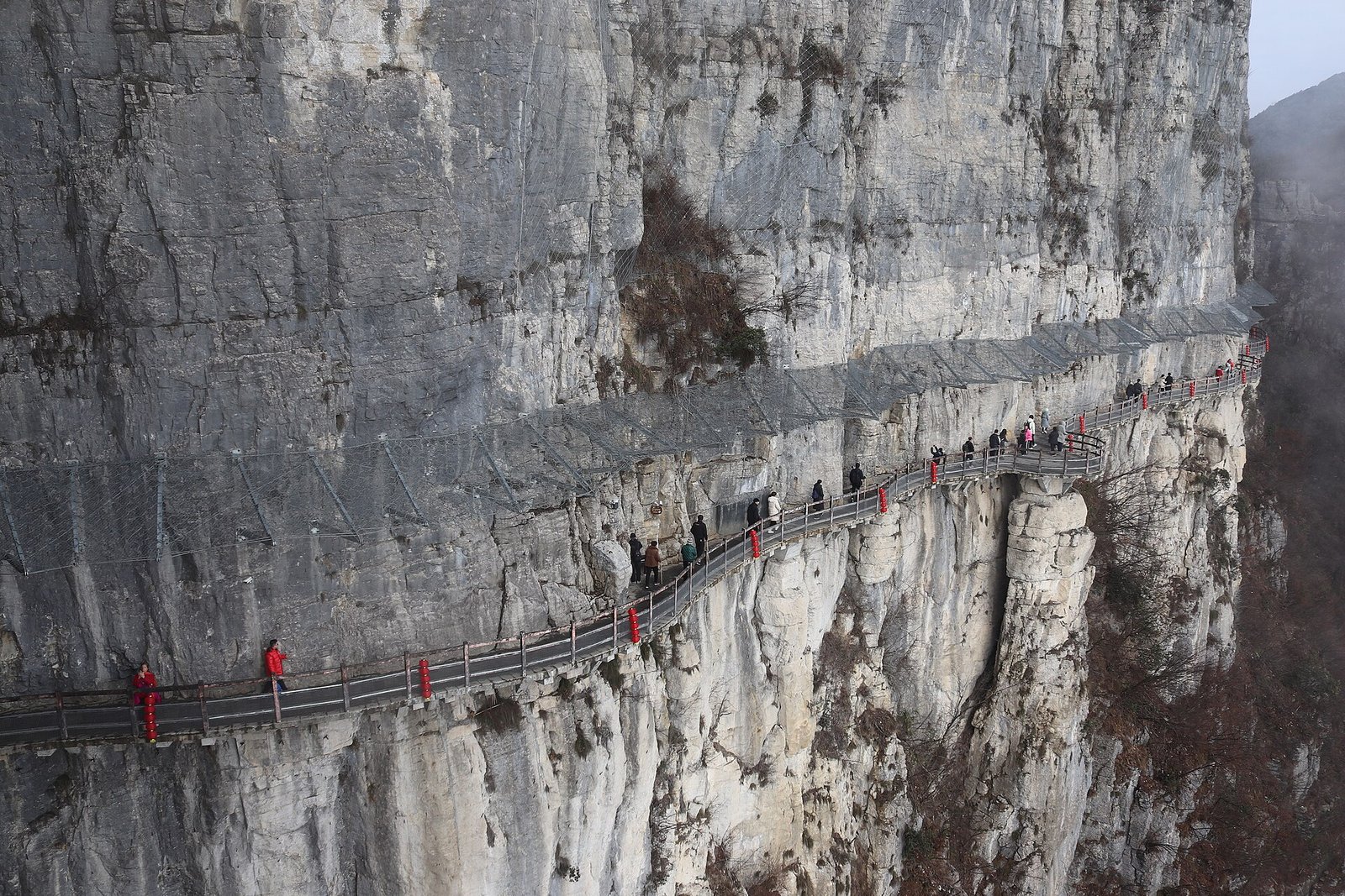 A concrete walkway pinned to a vertical cliff face at Qixingzhai in Enshi Grand Canyon, with two walkers in red visible against the rock