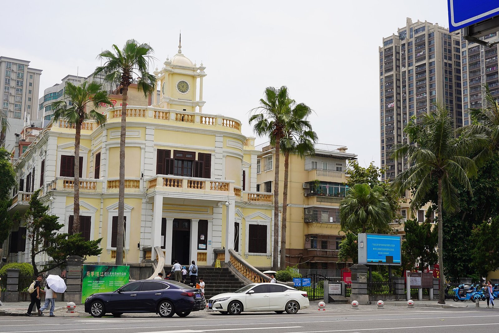 A pale yellow two-storey colonial building with a clock tower, balustraded balconies and a colonnaded entrance, with palm trees in front and a tall modern apartment block behind