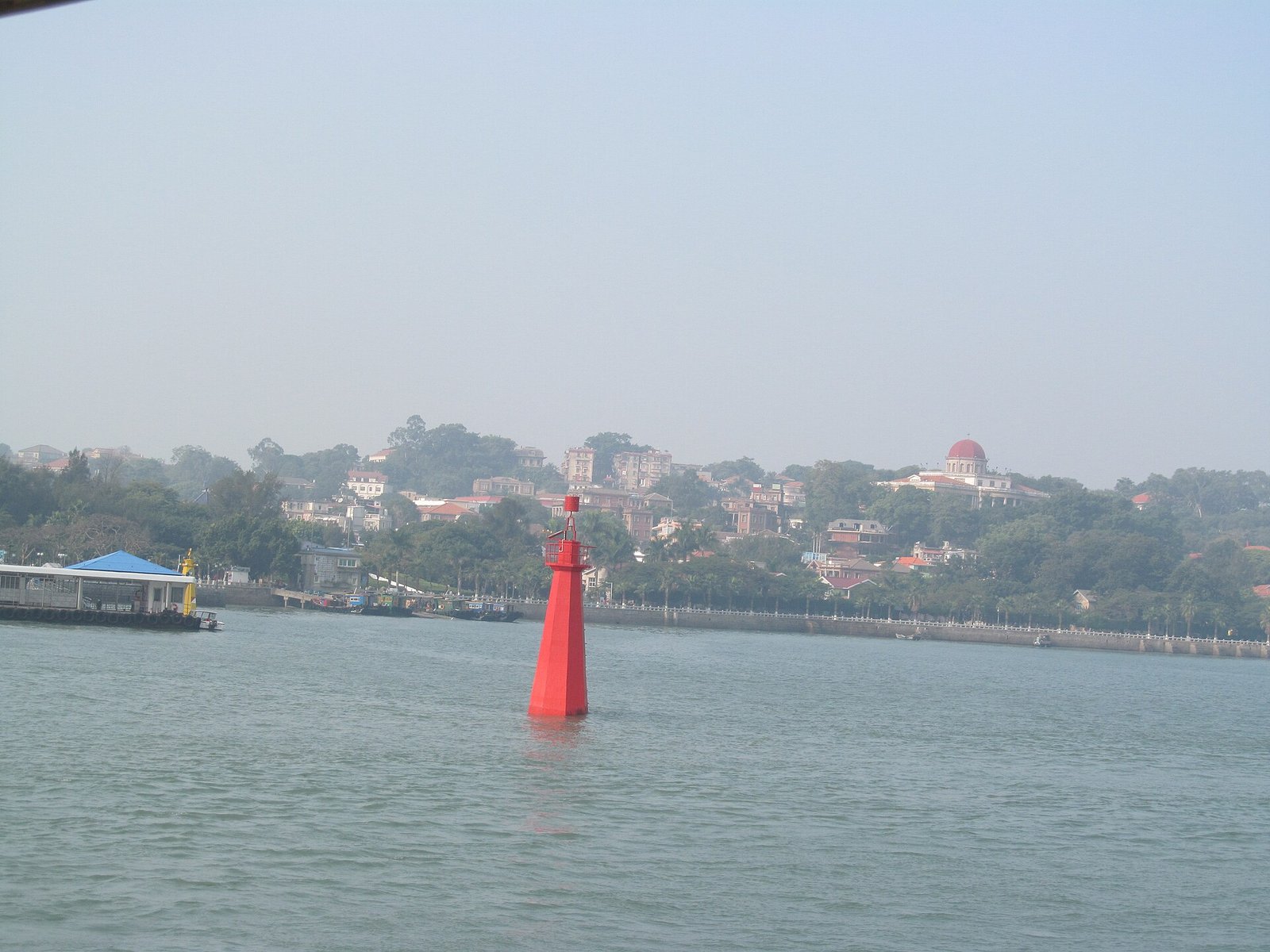 The wooded skyline of Gulangyu Island seen across the water from the ferry, with a red navigation beacon in the foreground