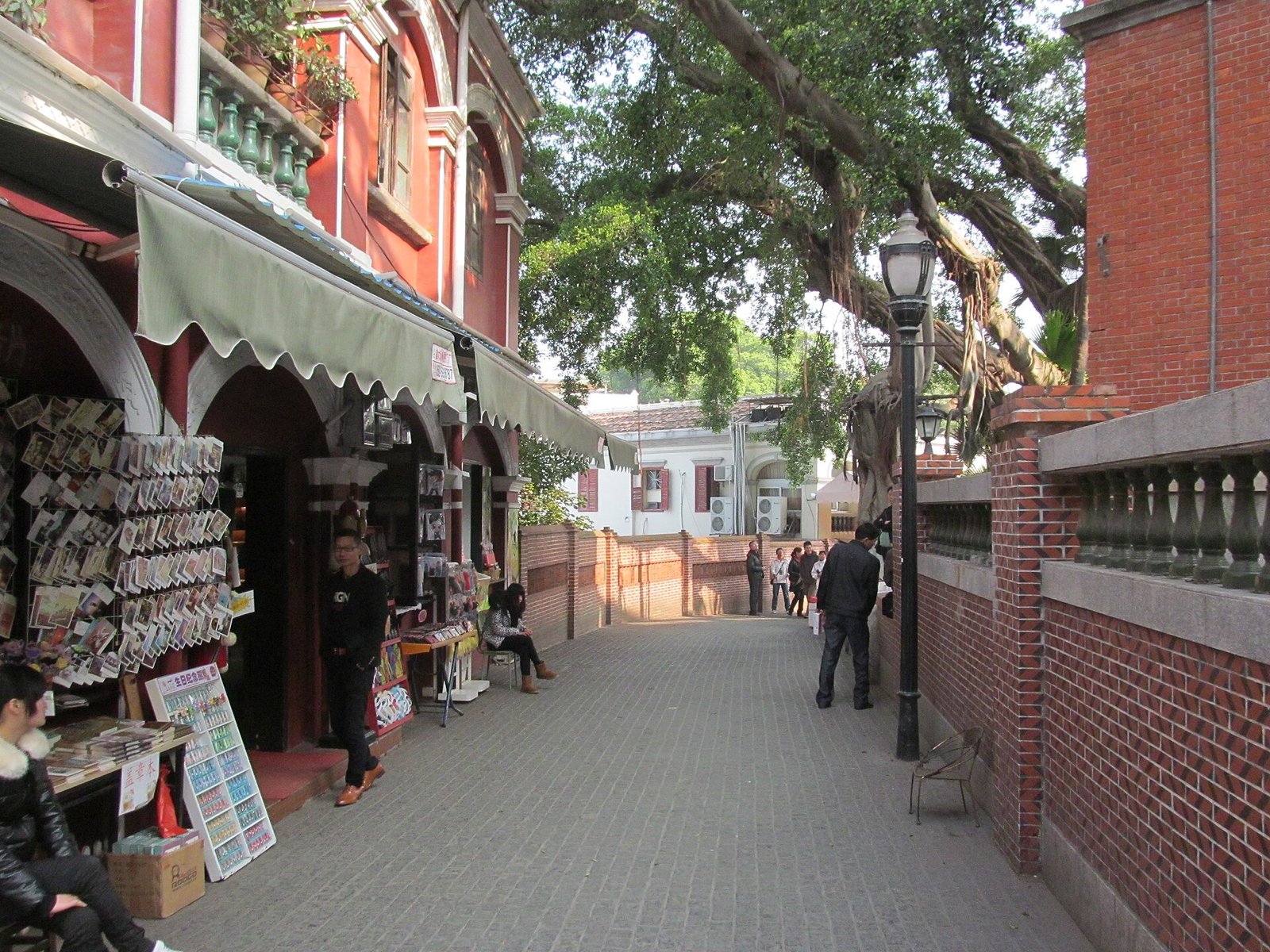 A paved lane on Gulangyu Island lined with colonial-era buildings, a book stall and plane trees, with a few visitors walking