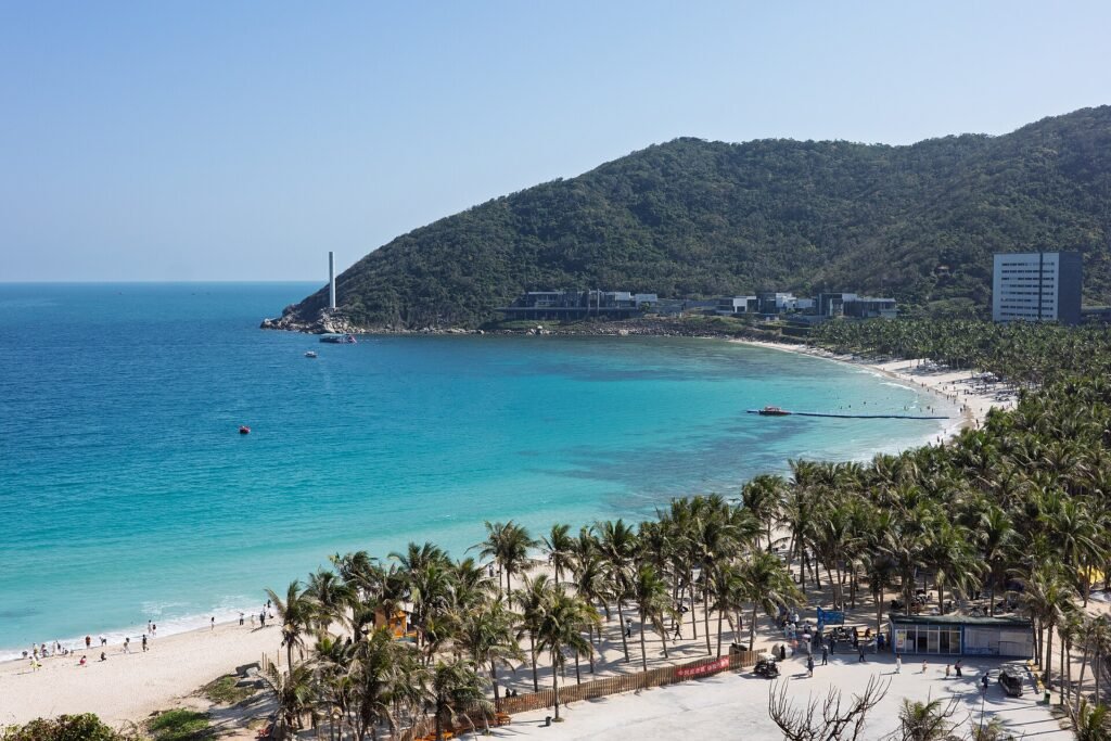 A curving turquoise bay with a forested headland, a road along the sand and resort buildings behind the trees