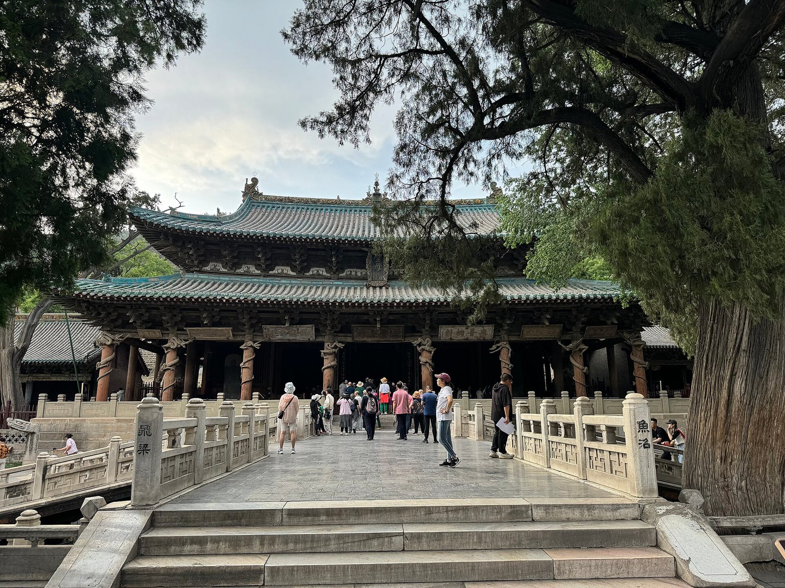 A large timber hall with a curved tiled roof behind a cross-shaped stone bridge on piers over a pool