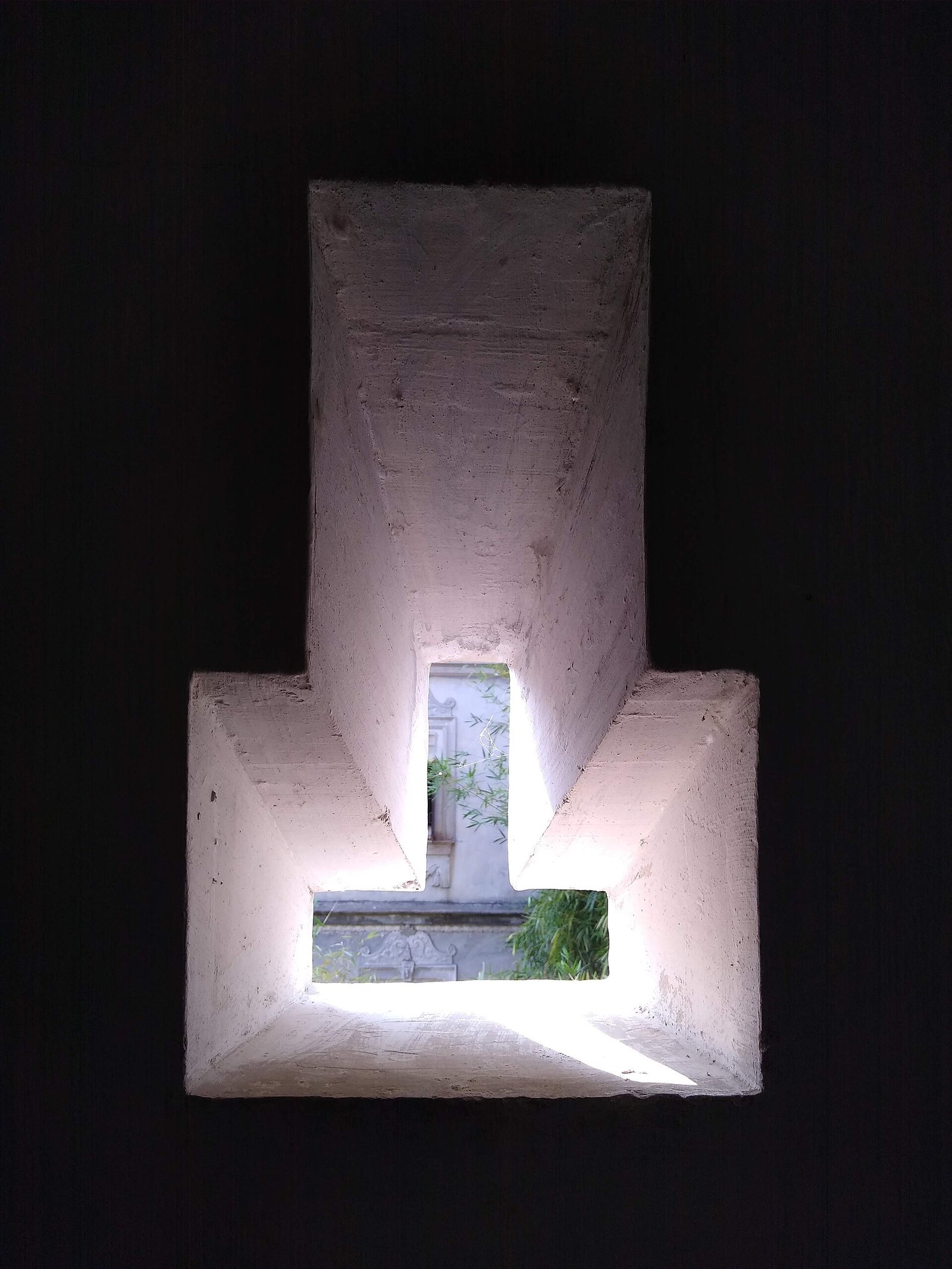 A narrow vertical firing slit set in a thick concrete wall, splayed inward, with a sunlit building and bamboo visible through it