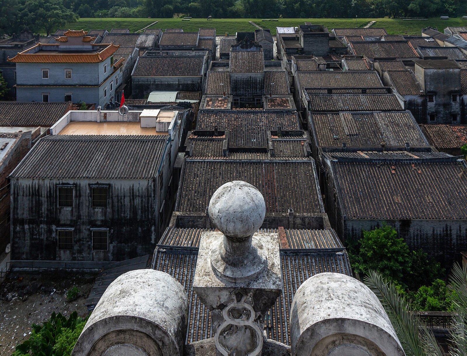 Grey tiled village roofs seen from the top of a tower, with a carved stone finial in the foreground and green fields beyond