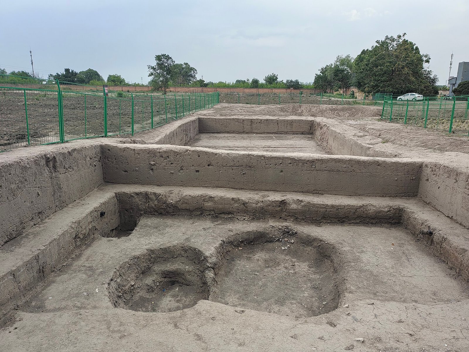 An archaeological excavation trench in farmland, showing rammed-earth foundation walls and two circular kiln pits, with green fencing and open fields beyond
