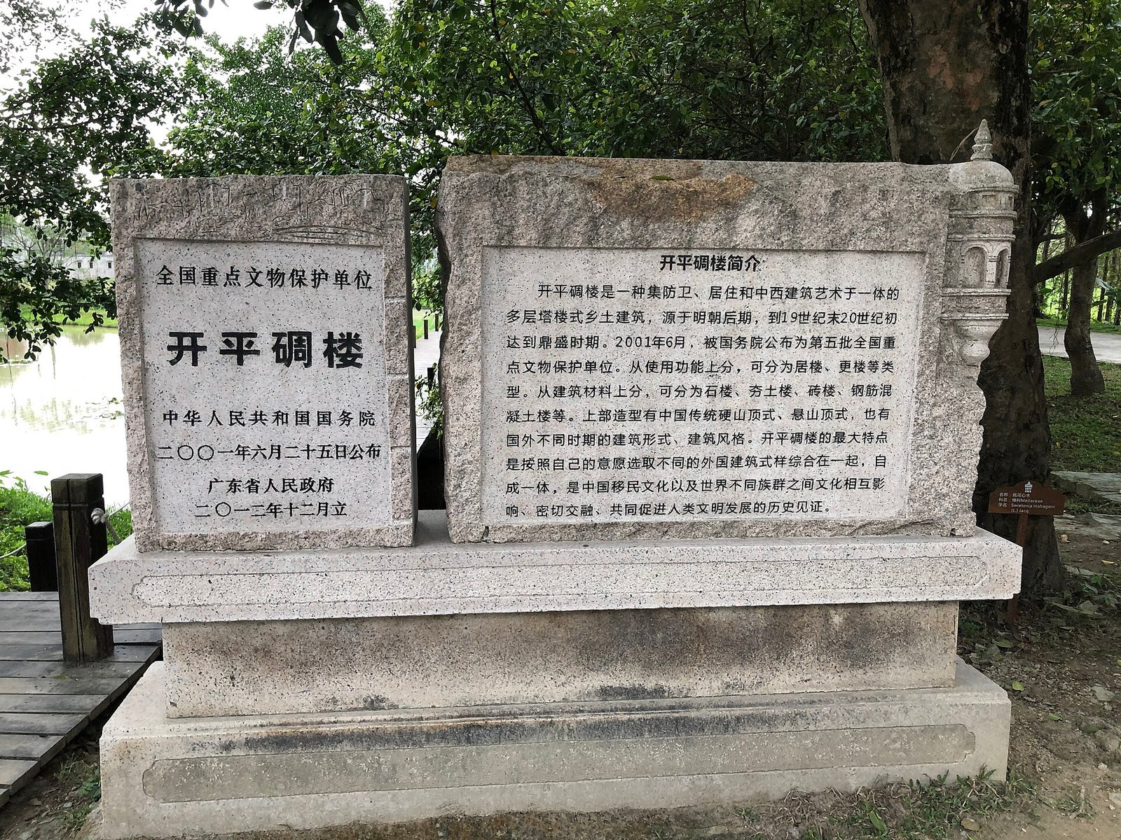 Two granite steles under trees, one reading Kaiping Diaolou and recording its 2001 national protection, the other carrying a description of the building type