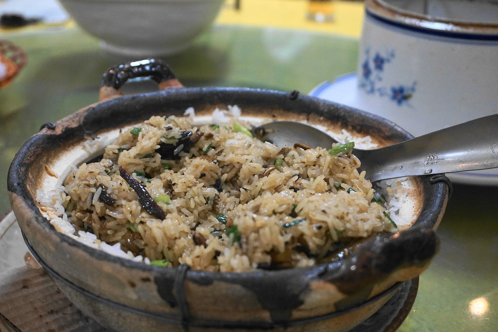 A clay pot of rice topped with shredded dark eel and spring onion, with a scorched crust visible at the edge
