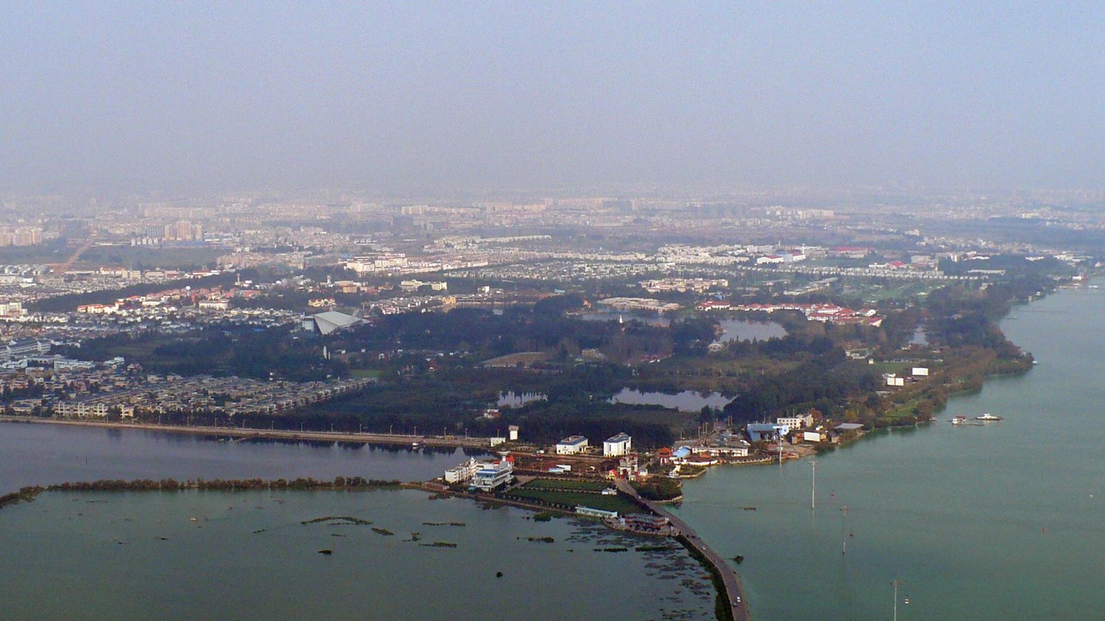 An aerial view of a large lake beside a city, with wooded shoreline, causeways and distant hills