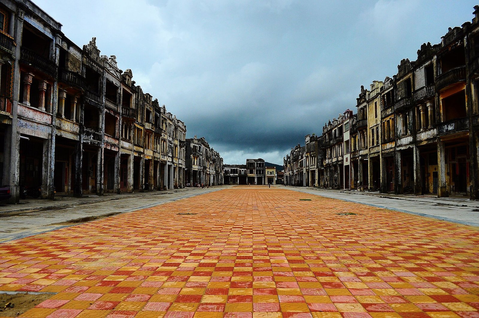 A long rectangular plaza paved in red brick, enclosed on both sides by three-storey arcaded shophouses with weathered facades, under a dark storm sky