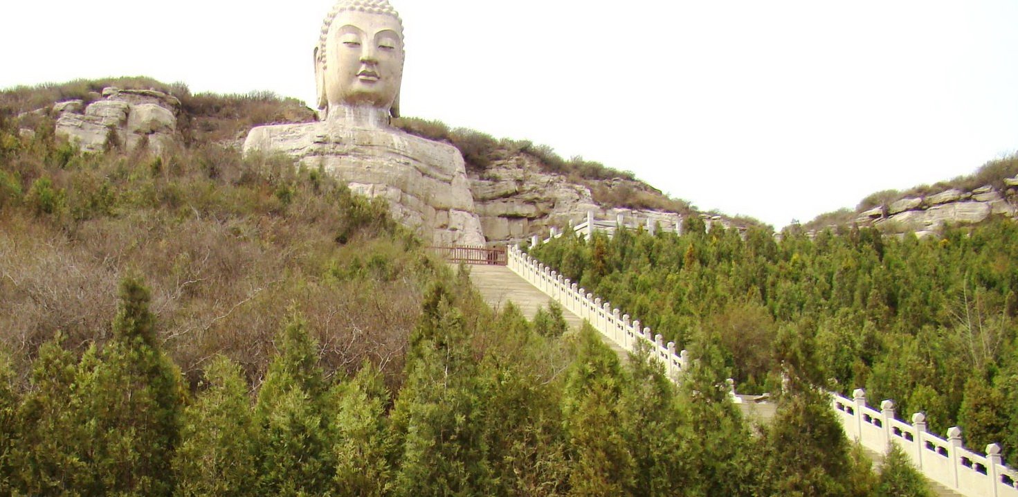 A very large seated Buddha carved into a cliff face, seen from below across a valley with trees on the slopes