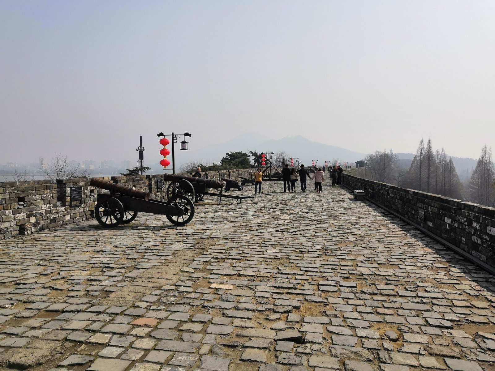 People walking along the paved top of an ancient city wall with cannons and distant hills