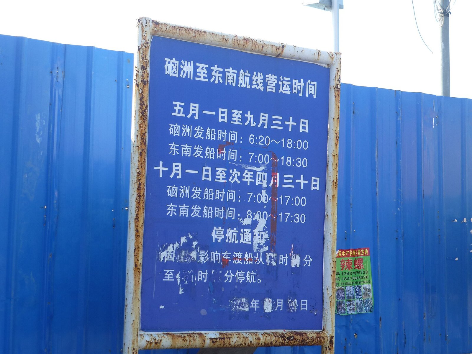 A weathered blue notice board bolted to a blue corrugated fence, hand-painted with ferry operating hours in Chinese and a handwritten line about typhoon suspensions