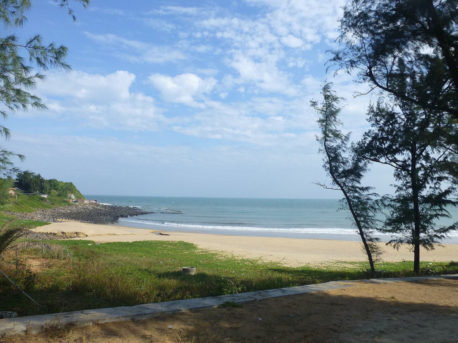 A pale sand beach curving between casuarina trees and a dark basalt and concrete sea wall, under a blue sky with scattered cloud