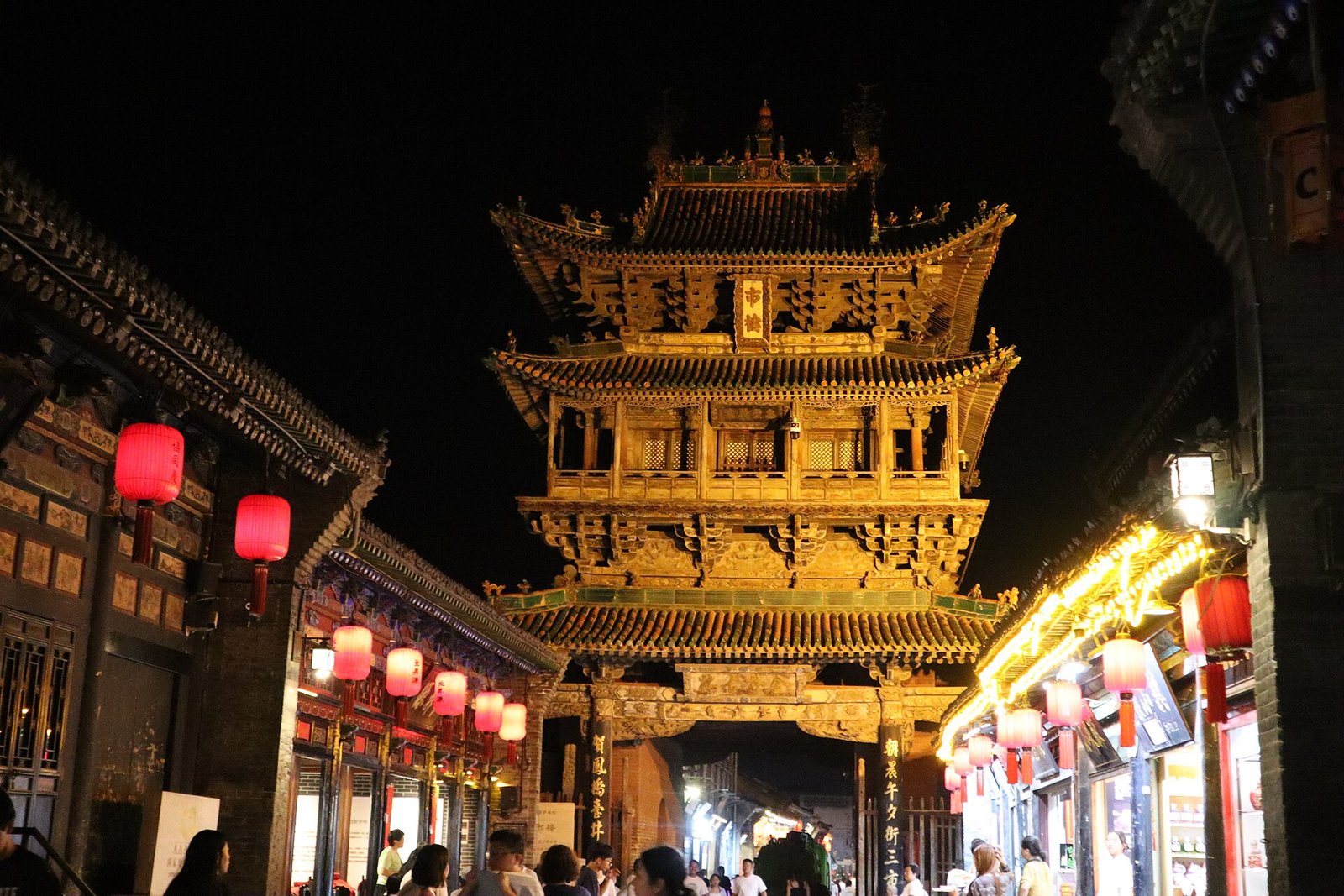 A multi-tiered wooden tower lit up at night in the old town of Pingyao, with red lanterns along the street