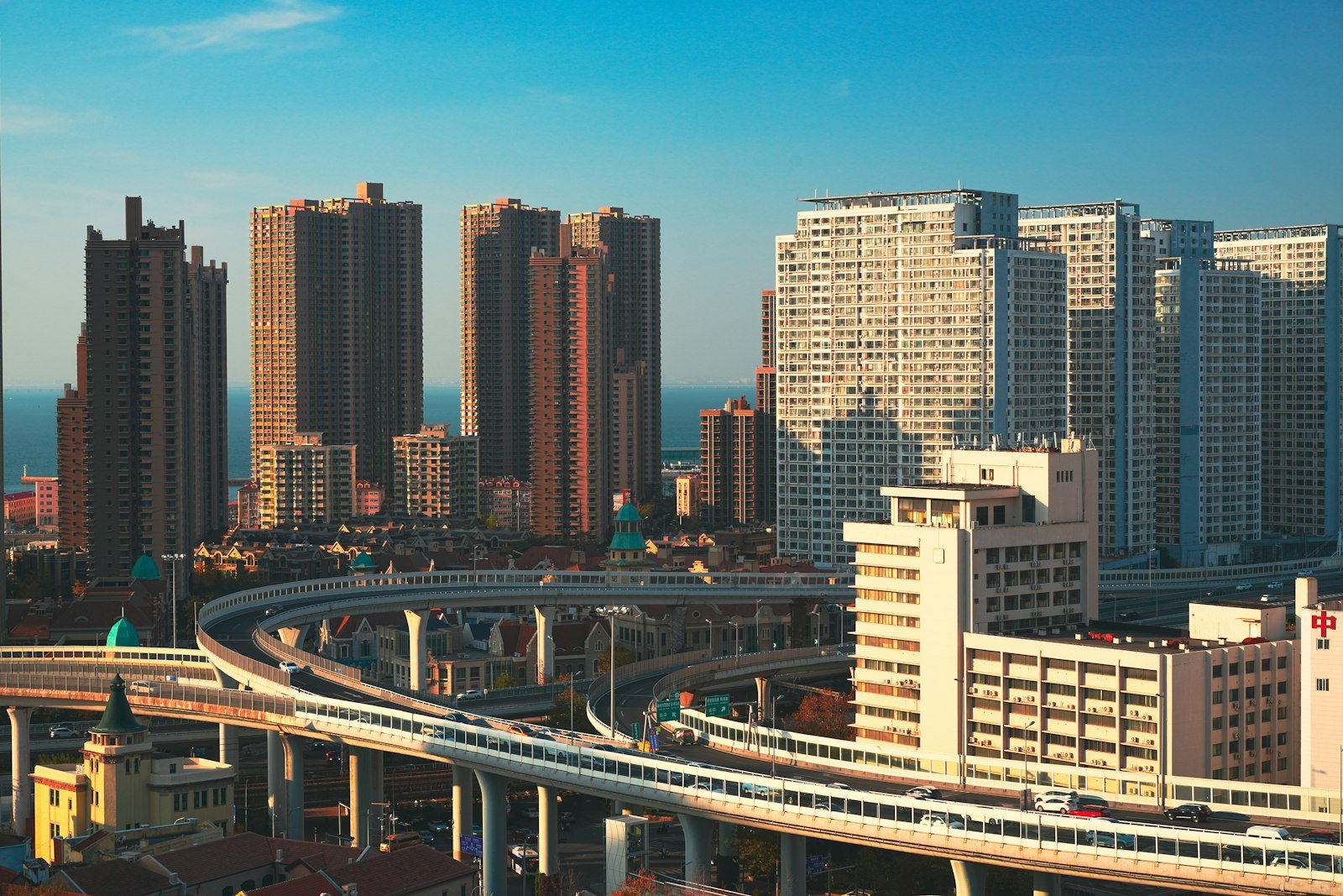 An elevated metro viaduct curving through Qingdao between high-rise towers and the sea