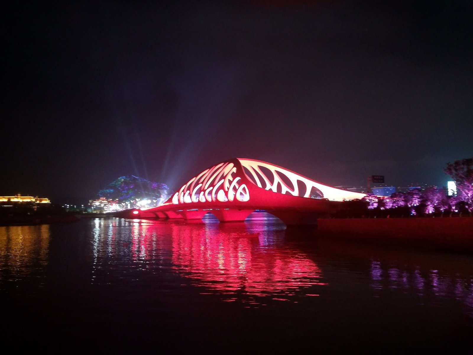 An arched footbridge lit red at night, its steelwork a lattice of leaf-shaped openings, reflected in still water