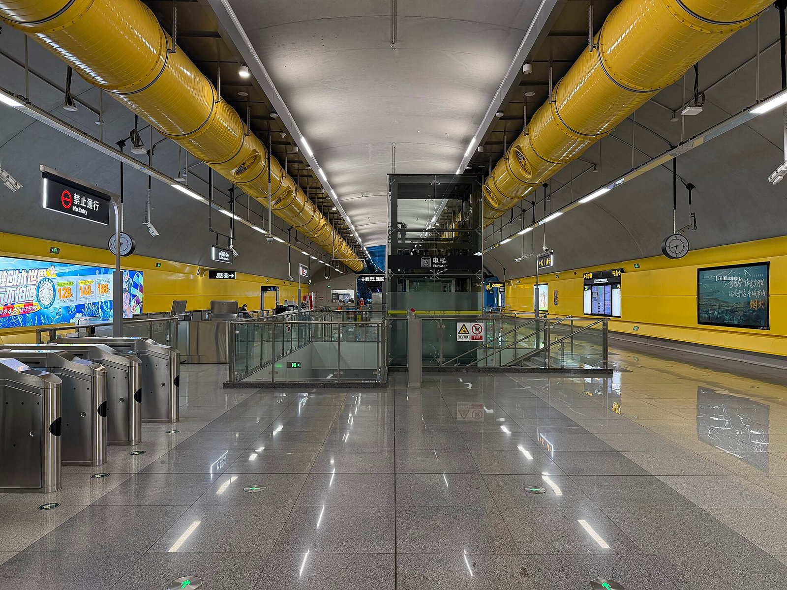 A wide metro concourse with a yellow slatted ceiling, fare gates, and a few passengers walking through