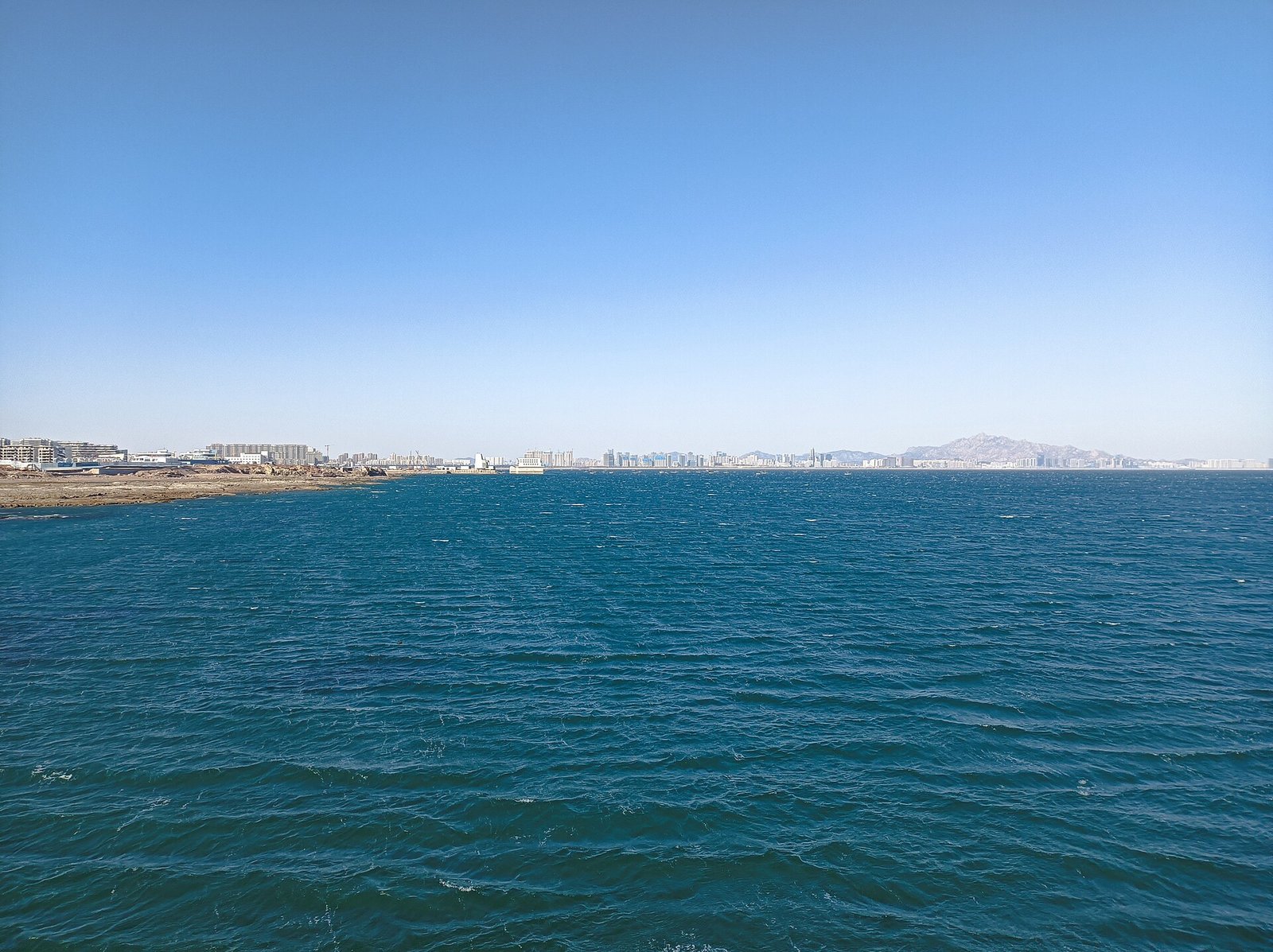 A calm blue sea meeting a low coastline, seen from the end of a pier under a pale sky