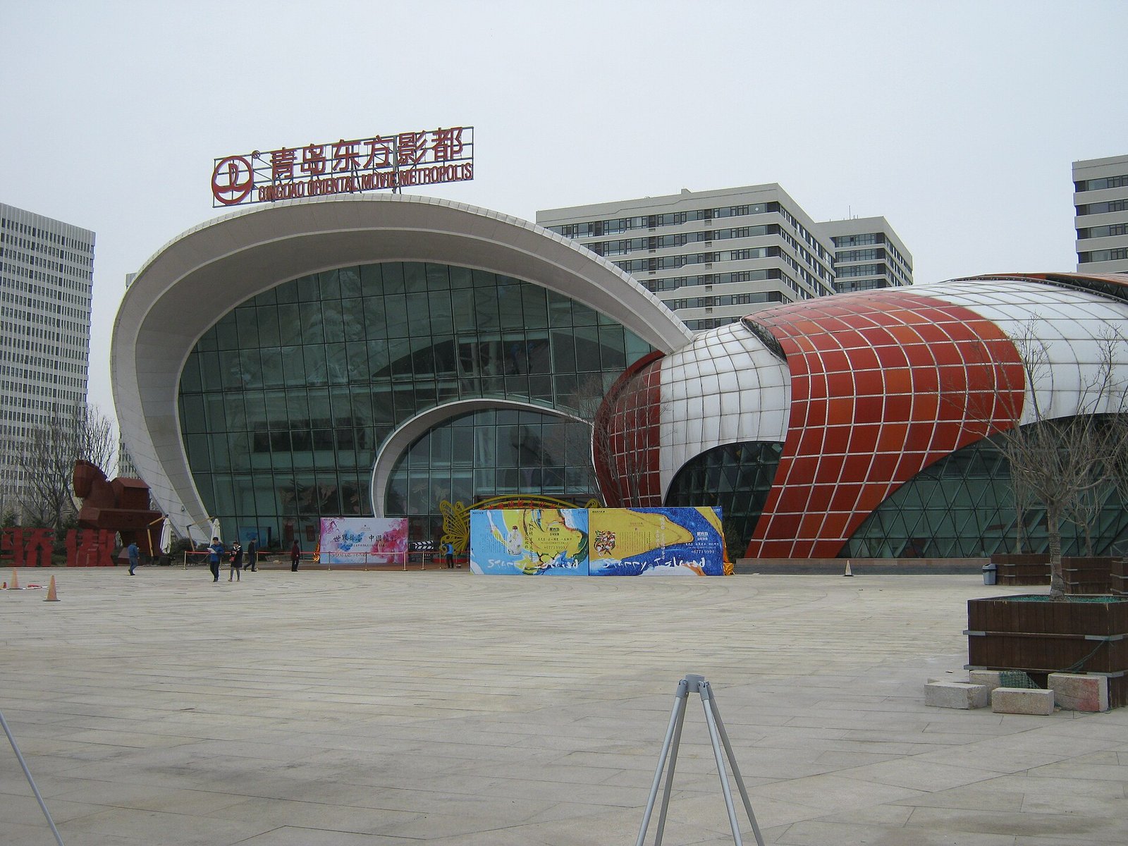 A wide modern gateway building with a curved white shell roof and a glass wall, a red and white striped drum beside it, and an empty paved plaza in front