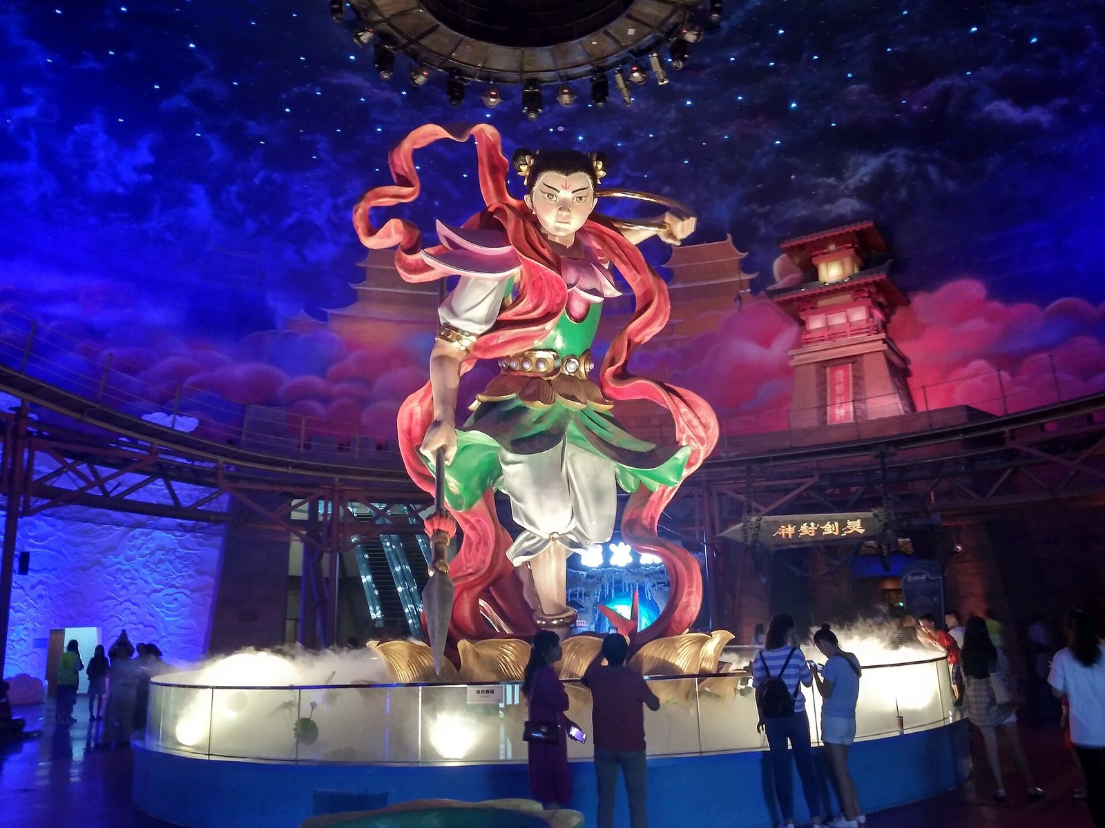 A giant painted figure with red ribbons inside a darkened hall, a ceiling painted as a starry sky with pink clouds, theatrical fog at the base, and visitors photographing it from a railing