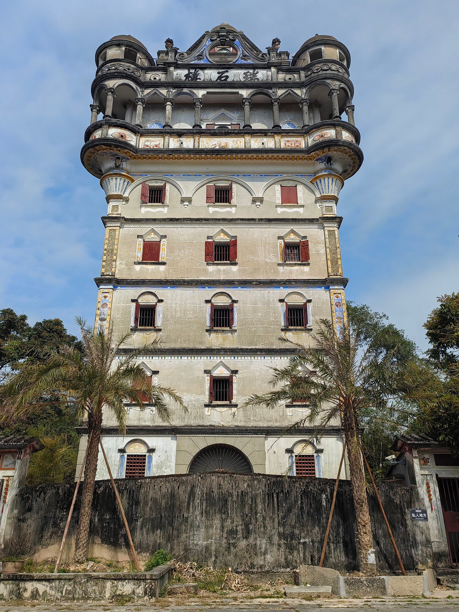 A nine-storey concrete tower with balconies, columns and a pavilion at the top, standing above a village roofscape