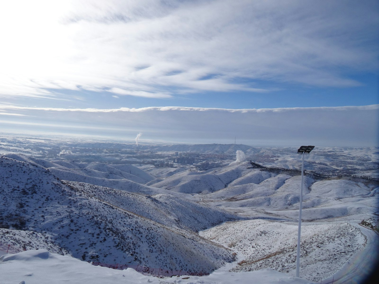 A ski lift tower standing on a snow-covered slope above the snow line, with a ski area and valley beyond