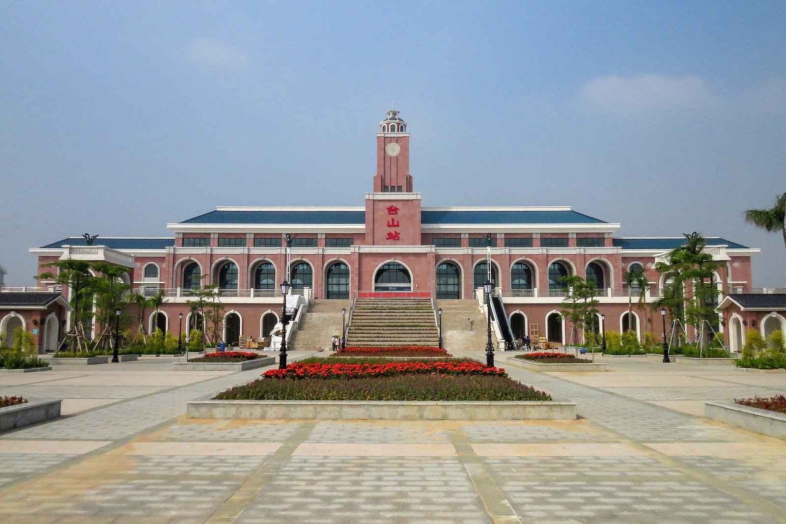 A modern station building with a clock-tower-like structure on the left and a wide paved forecourt
