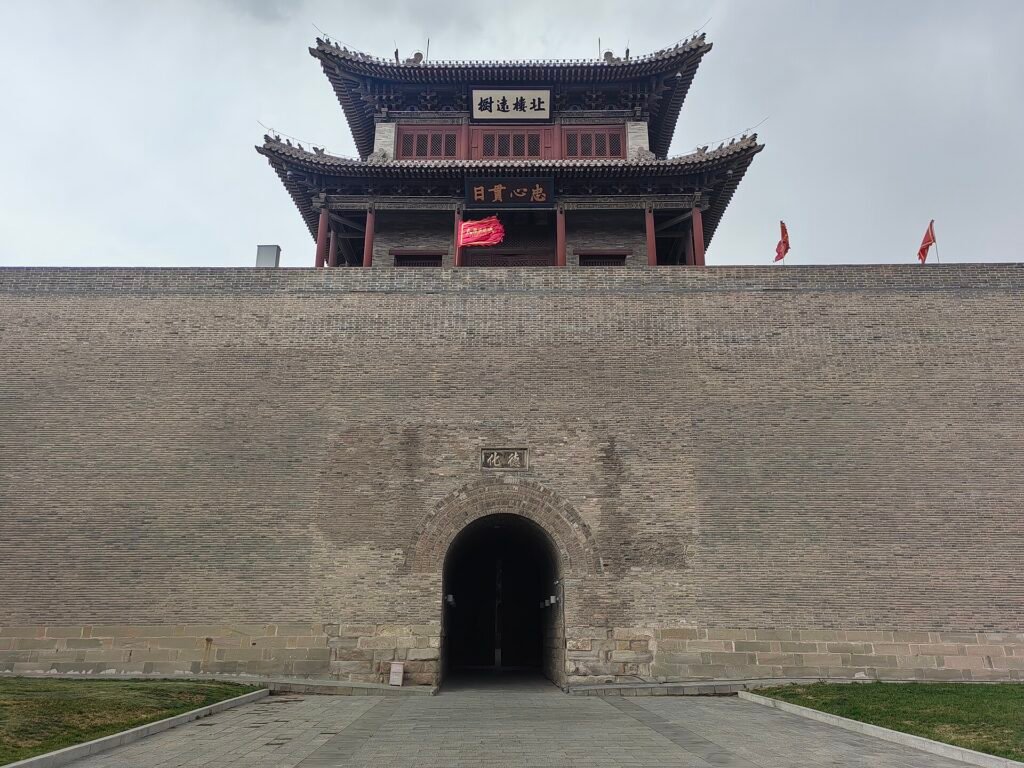 A grey brick city gate with a double-eaved timber tower above a dark archway, red flags on the parapet and young grass at the base