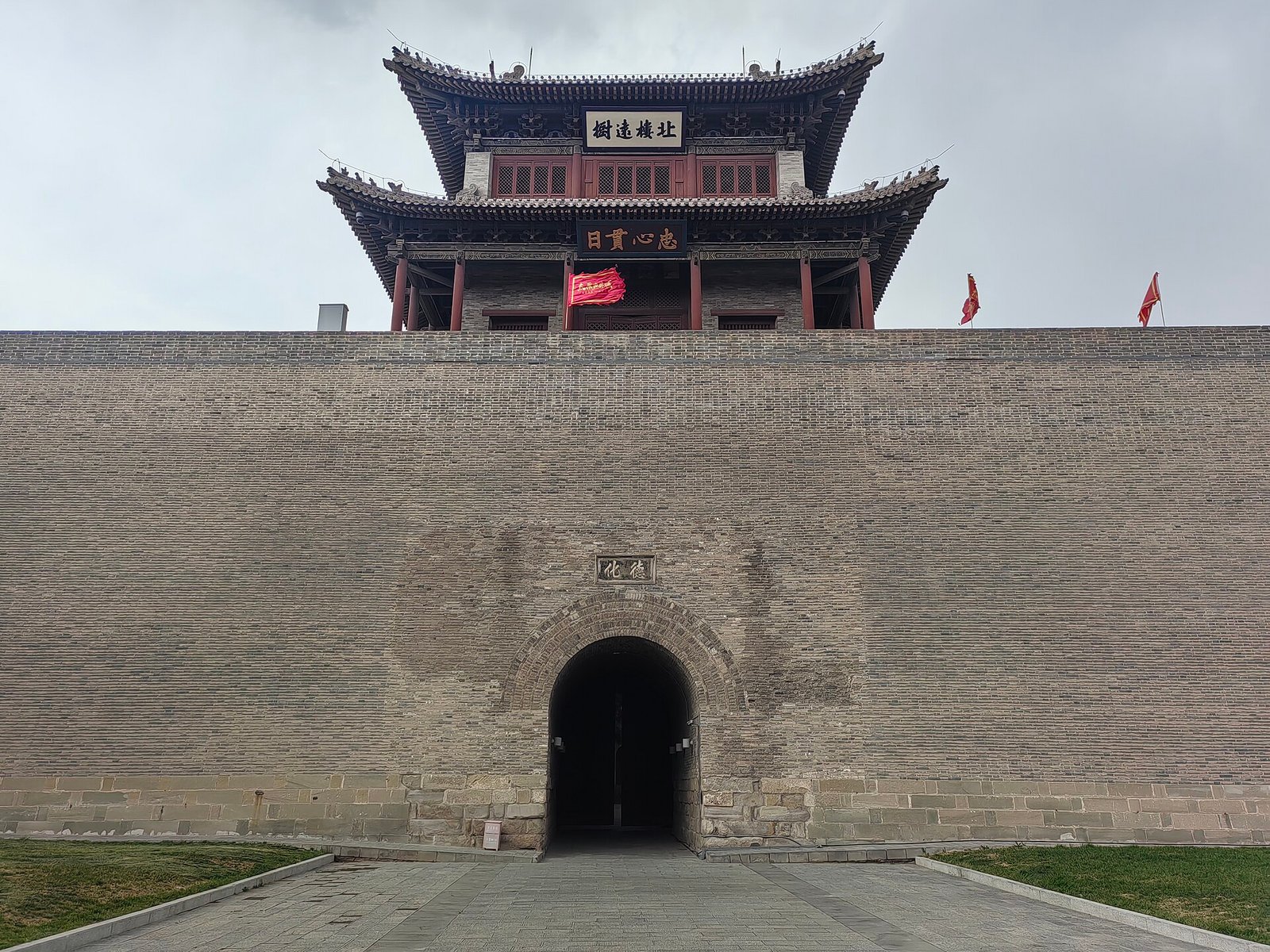 A grey brick city gate with a double-eaved timber tower above a dark archway, red flags on the parapet and young grass at the base