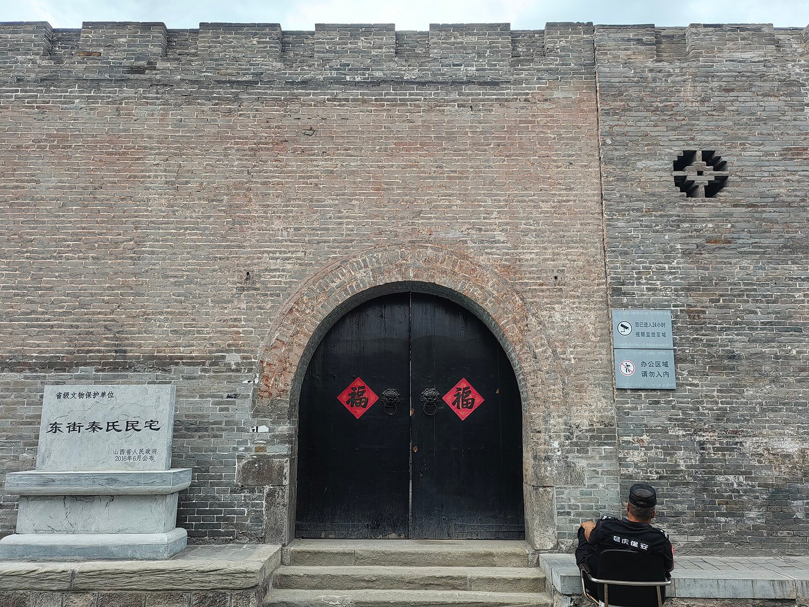A grey brick courtyard house with a tiled gate, a stone plaque beside the door and a person standing at the threshold