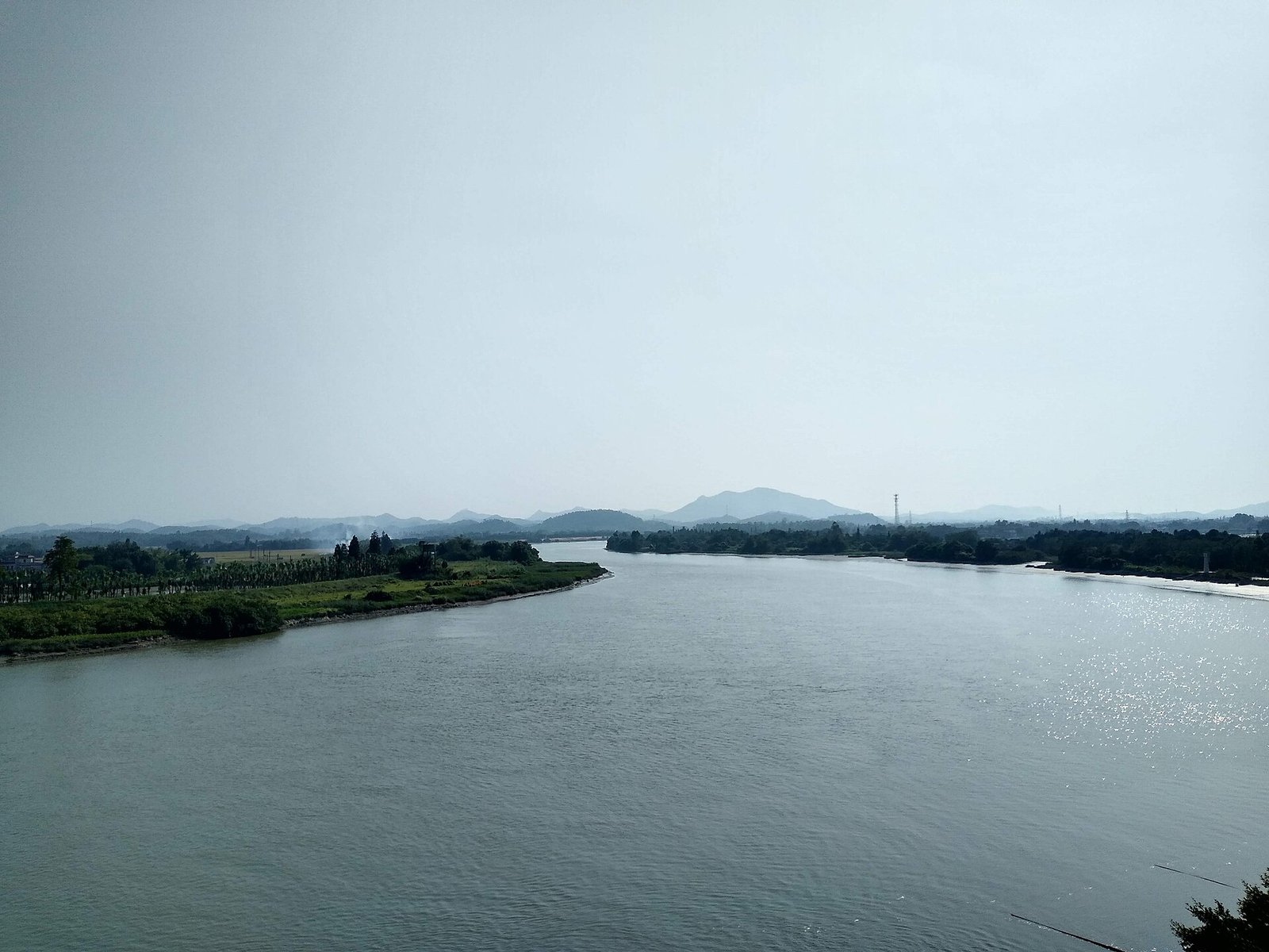 A wide green river curving between low wooded banks under a hazy sky, with hills on the horizon