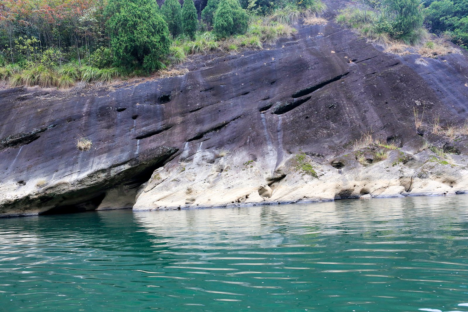 A sheer red sandstone cliff cut with caves rising directly out of the green water of the Nine-Bend Stream