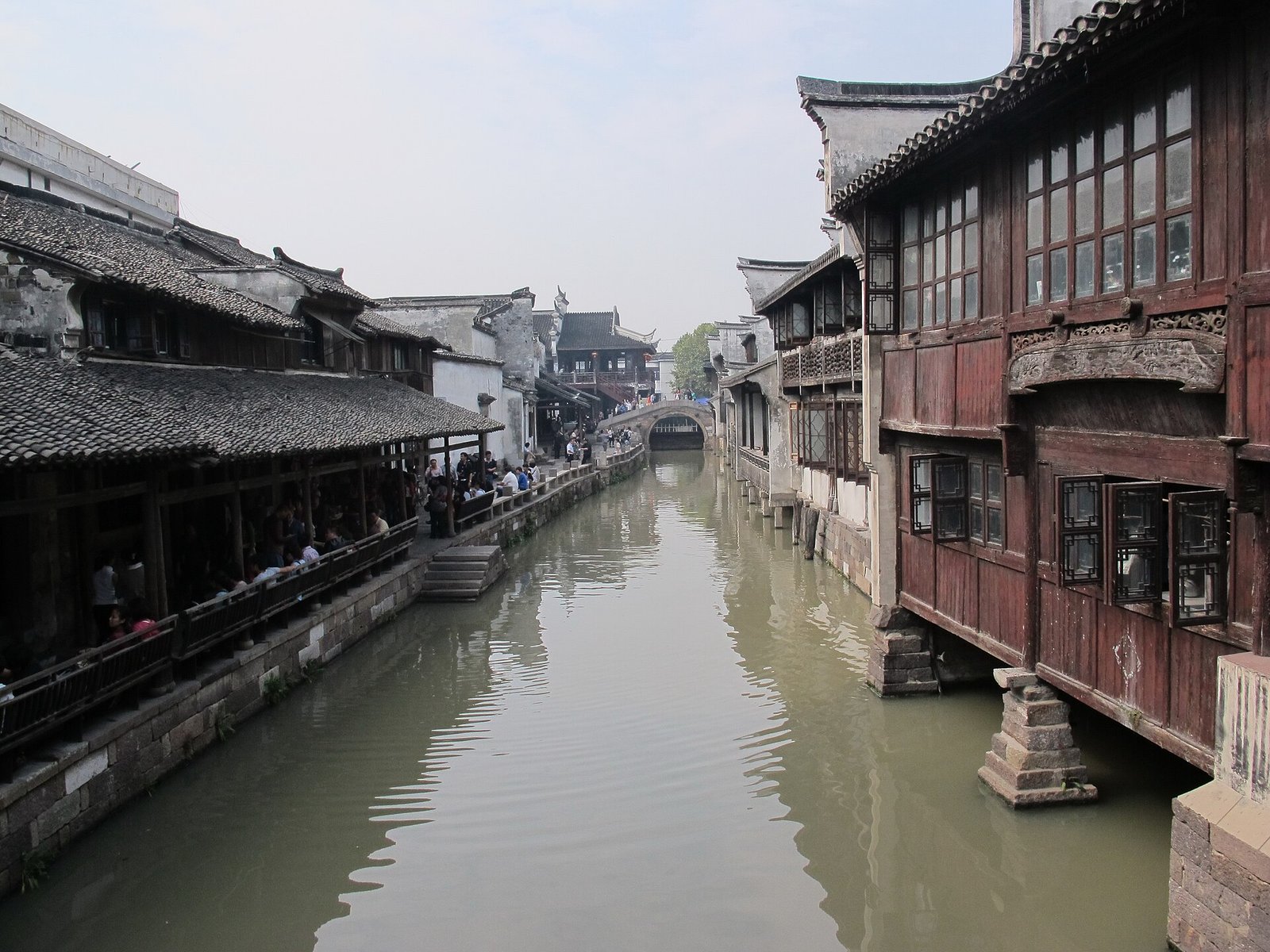 A canal in Wuzhen Dongzha lined with white-walled houses and dark tiled roofs, with a stone bridge in the middle distance