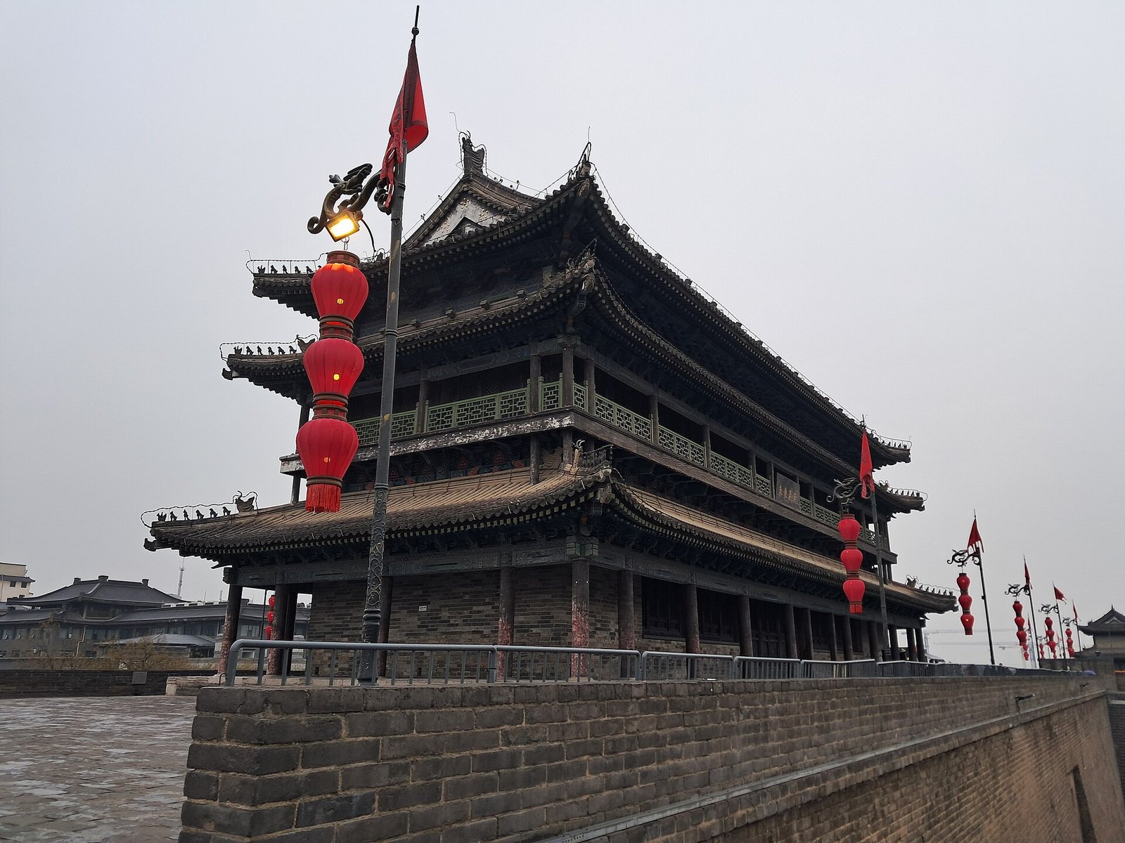 A three-tiered Chinese gate tower of grey brick and dark timber, hung with red lanterns, standing on a city wall
