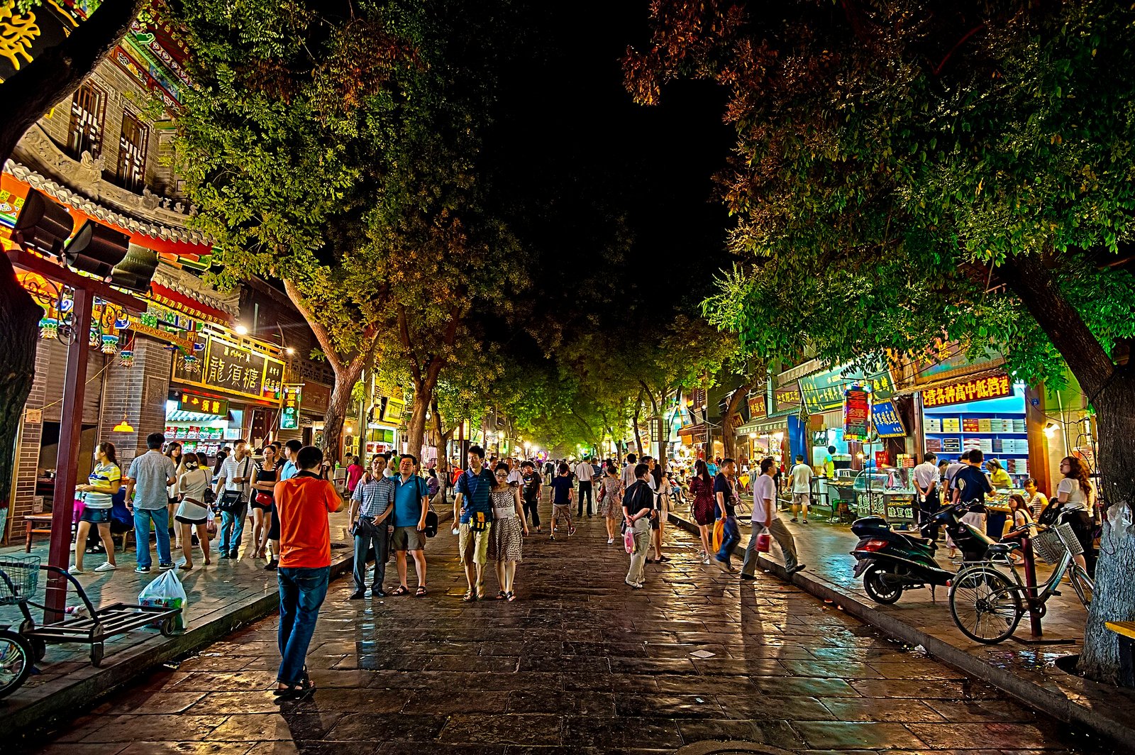 A crowded Chinese food street at night under trees, with lit shopfronts on both sides
