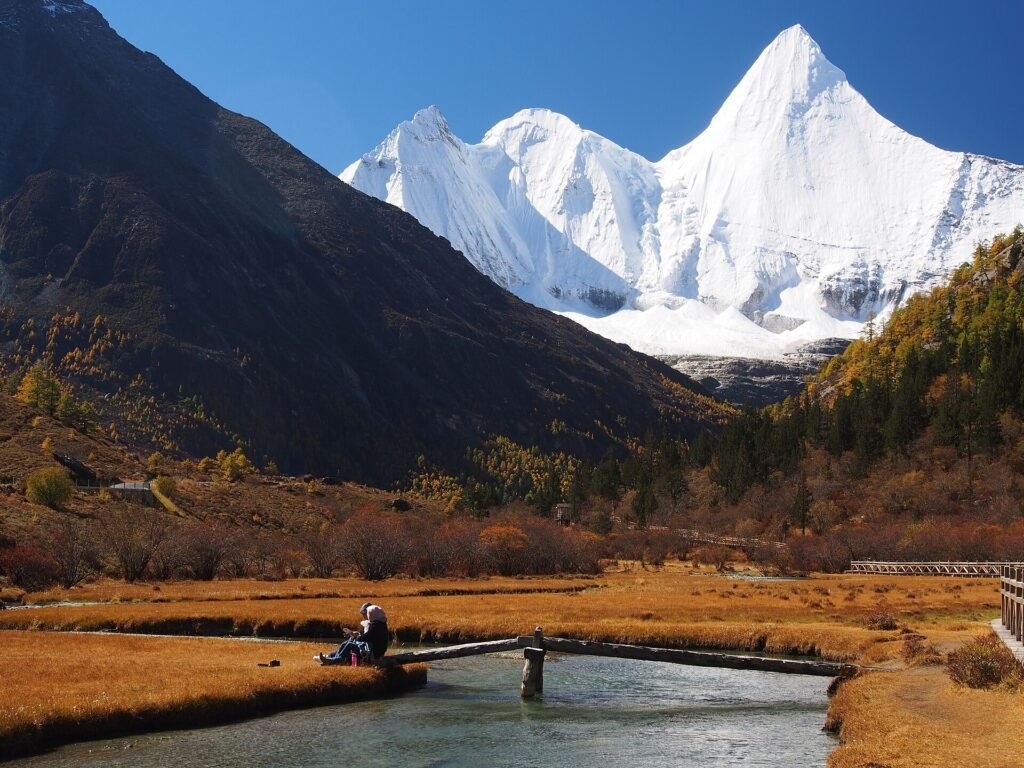 The sharp snow pyramid of Jampelyang rising above a golden autumn meadow with a small pool reflecting it