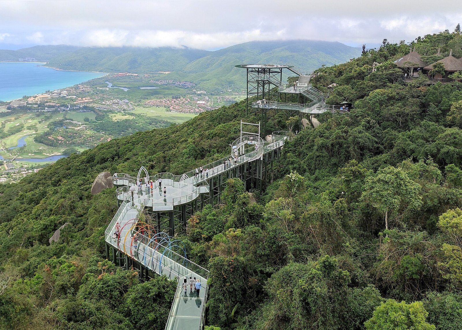 A long steel and glass walkway running along a ridge above forest, with hills and sea beyond