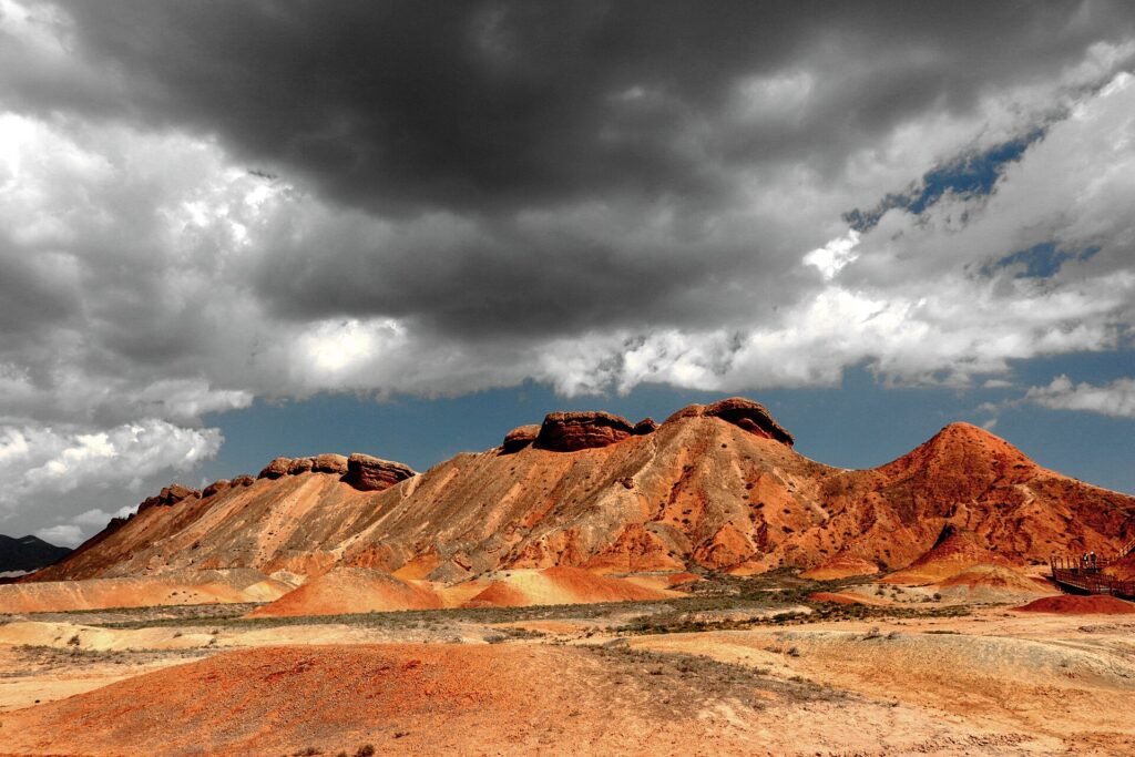 Banded red and ochre hills at Zhangye Danxia under a dark storm sky, with a section of boardwalk at the right edge