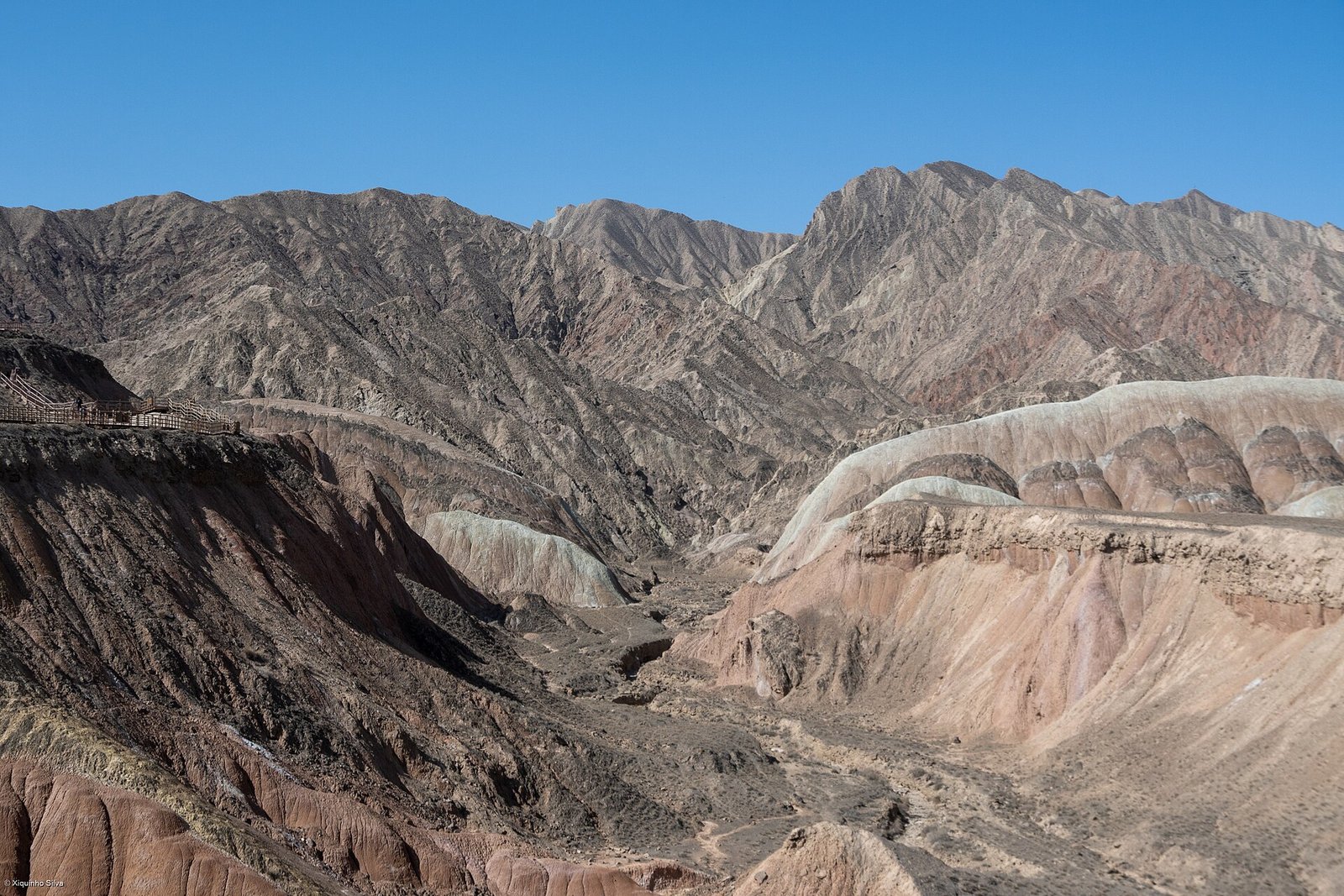 A broad view of the coloured hills at Zhangye Danxia with grey gravel terraces in the foreground and mountains on the horizon