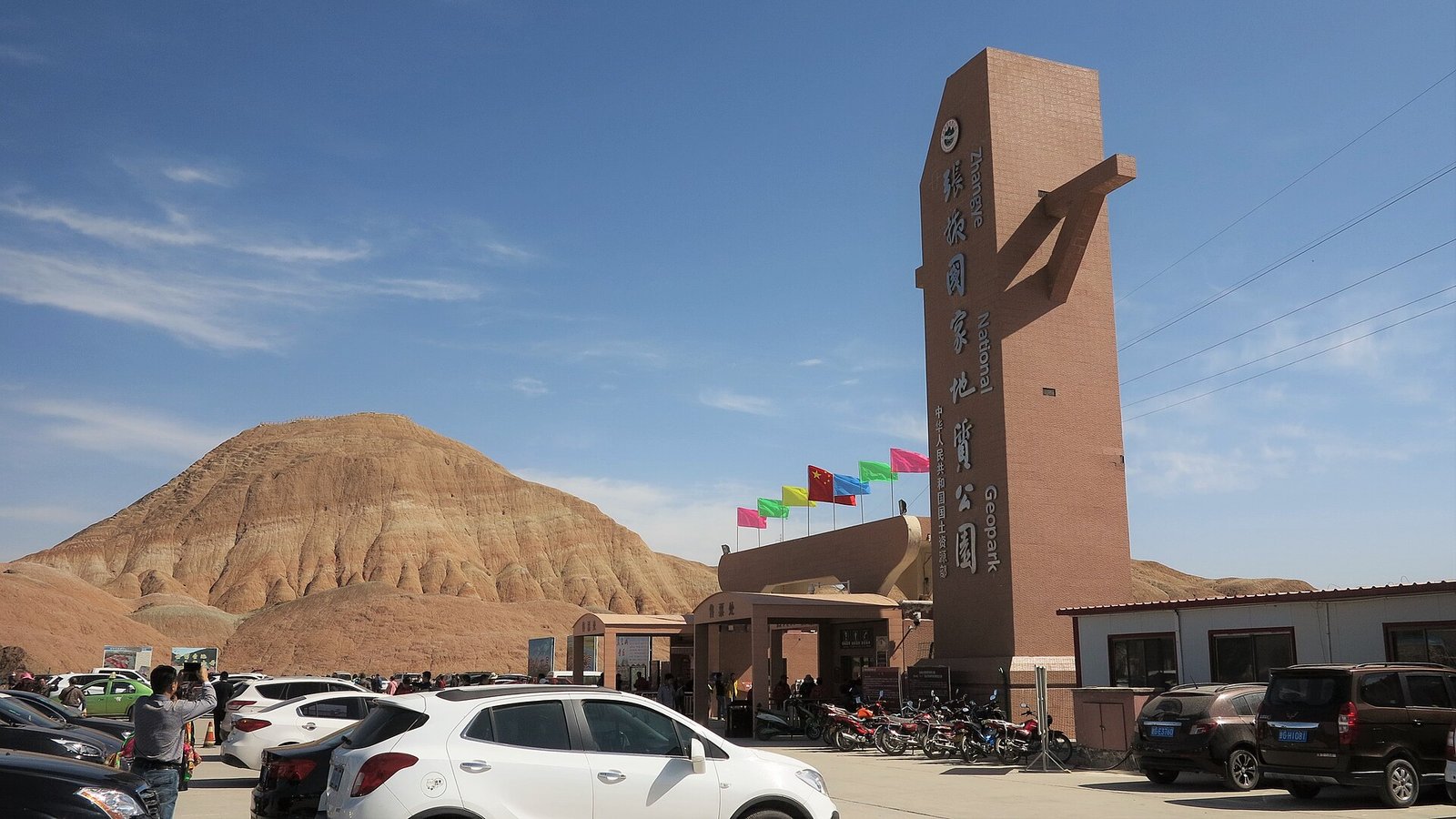 The entrance pillar at the north gate of the Zhangye Danxia scenic area, lettered in English and Chinese as Zhangye National Geopark, with a car park in front