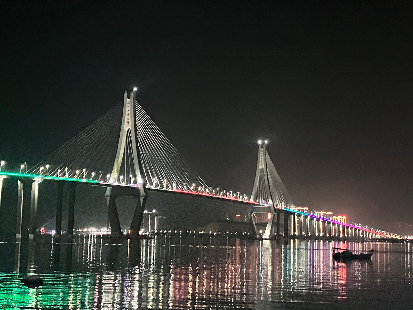 A cable-stayed bridge with two tall pylons lit in colour at night, reflected in flat water, with a small boat in the foreground
