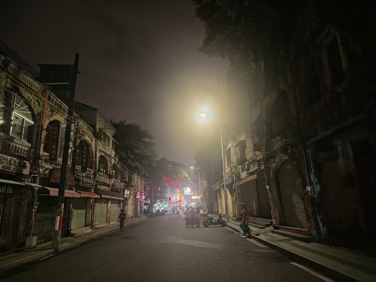 A narrow street at night lined with two-storey arcaded shophouses, lit by a single street lamp in humid haze, with closed roller shutters and a few people walking